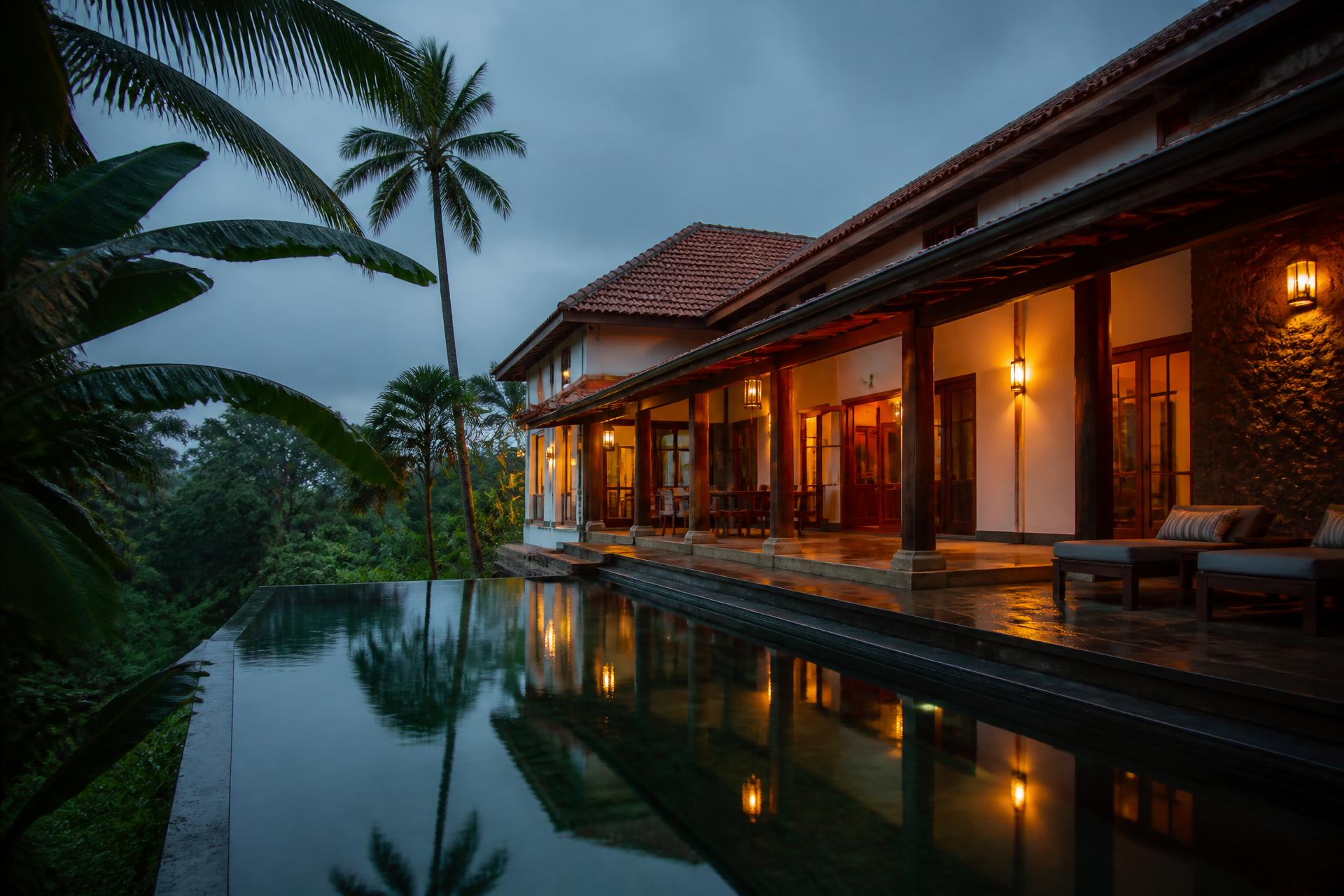 A Goan villa at dusk, its verandah lit warm above a still pool, palms heavy with monsoon rain